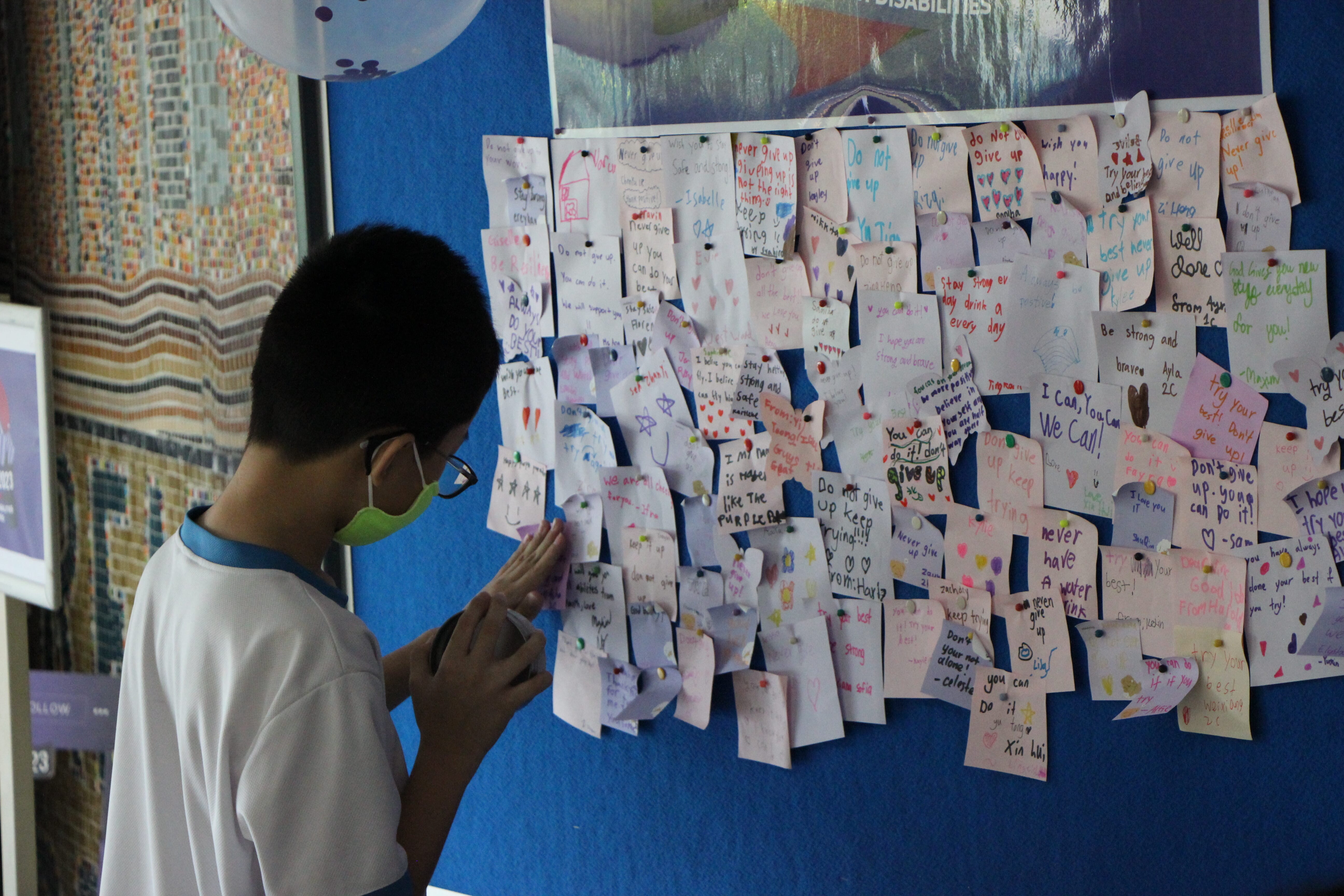 A masked boy in a white shirt reads handwritten notes of encouragement pinned to a blue board.
